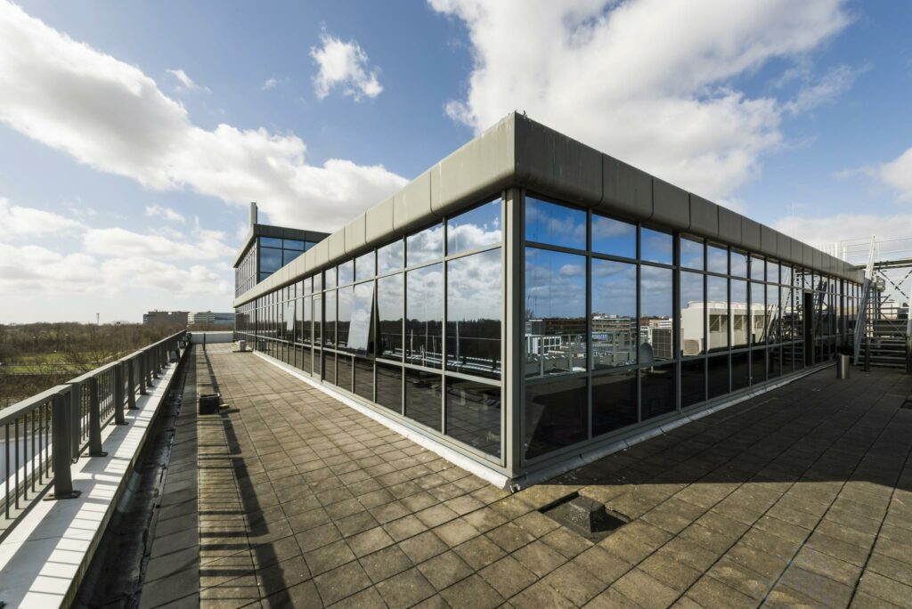 Dakterras met grote glazen gevel van een modern kantoorpand aan de Anthony Fokkerweg onder een deels bewolkte blauwe lucht.